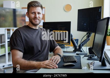 Portrait of smiling software developer looking at camera while sitting at desk with computers Stock Photo