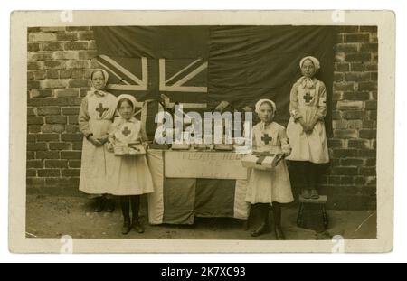 Original and charming WW1 era postcard of working class children dressed in red cross uniforms, with a stall raffling or selling their toys and games, sweets and potted plants to help raise money for wounded soldiers, Union Jack flag in background, 1914-1918 U.K. Stock Photo