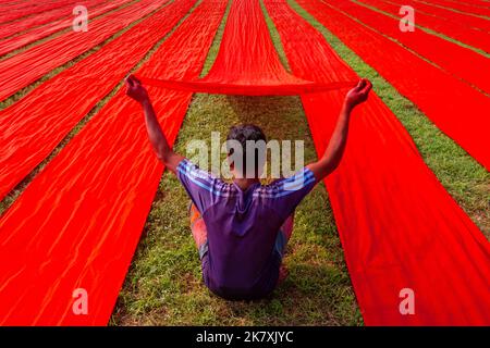 Narsingdi, Dhaka, Bangladesh. 19th Oct, 2022. Hundreds of meters of bright red fabrics are laid out in neat rows across a field in Narsingdi, Bangladesh. Known as ''Lal Shalu'' to the locals, the long red cloths are set out to dry under the hot sun, having been dyed with bright red color. The use of sunlight to dry out the fabrics reduces production costs as it is cheaper and more sustainable. The eco-friendly drying method spans an area equal to 5 football fields and takes up to 6 hours to complete after being placed by workers at sunrise. The cloth, which sells for less than 10 GBP, is use Stock Photo