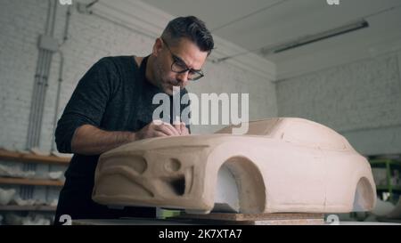 A ceramist works closely on the design details of an unfinished eco friendly electric car prototype sculpture made of raw clay. Designer wearing glasses and an apron works in a ceramic workshop. Stock Photo