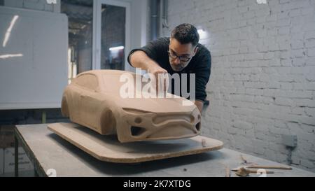Senior automotive designer sculpts the prototype model of the car made of clay using sculpting tools. Experienced designer works in a workshop. Automotie industry. Stock Photo