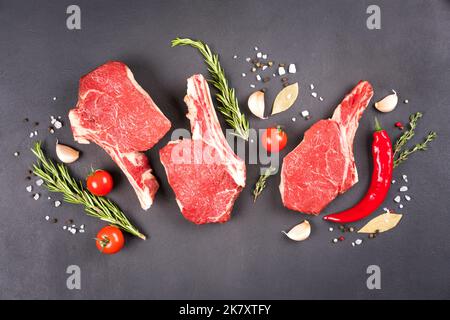 Raw ribeye beef steaks with seasonings over dark concrete background. Cowboy steak on bone ready for grilling. View above Stock Photo