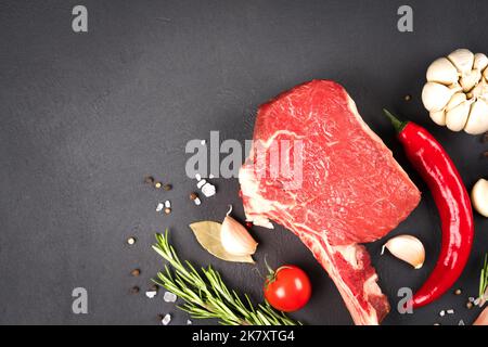 Raw ribeye beef steaks with seasonings and vegetables over dark concrete background with copy space. Cowboy steak ready for grilling Stock Photo