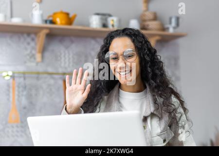 Close up of happy hispanic woman talking on video call at home, smiling and waving greeting gesture using laptop for remote communication sitting in kitchen. Stock Photo