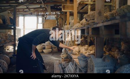A ceramist wearing an apron and black framed glasses places his art work in a clay pottery studio. Ceramic workshop to showcase his art work. Beautiful hand made clay sculptures. Stock Photo