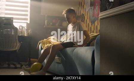 A teenager boy plays football while sitting on the bed alone in his room. Balancing the ball on his finger. Performing tricks. Sports, hobbies concept. Stock Photo