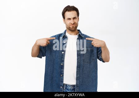 Skeptical guy pointing at himself with upset frowned face, being unconfident, showing doubt, standing over white background Stock Photo