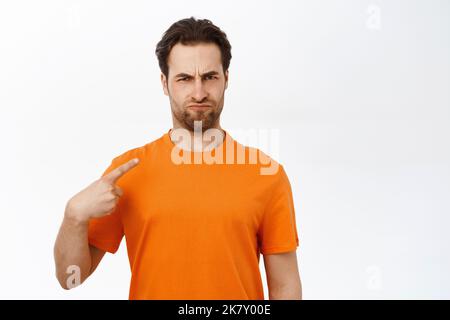 Portrait of man with angry face pointing at himself, looking grumpy, standing over white background Stock Photo