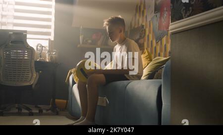 A teenager boy plays football while sitting on the bed alone in his room. Balancing the ball on his finger. Performing tricks. Sports, hobbies concept. Stock Photo
