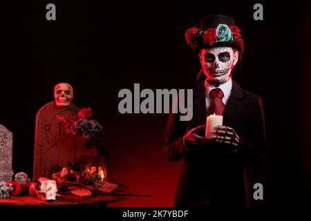 Young woman with skull make up holding candle burning in studio, being dressed as santa muerte to celebrate dios de los muertos. Looking like mexican goddess of death on day of the dead. Stock Photo