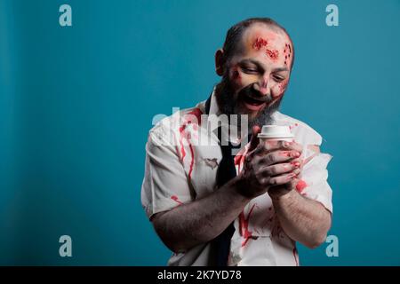 Horror demon looking at cup of coffee, wanting to dirnk beverage and being aggressive in studio. Scary evil monster holding cup and having bloody wounds, apocalyptic eerie devil. Stock Photo