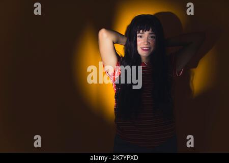 Young brunette white woman standing in yellow spotlight in relaxed pose with hands behind head looking at camera smiling. Horizontal studio shot. High quality photo Stock Photo