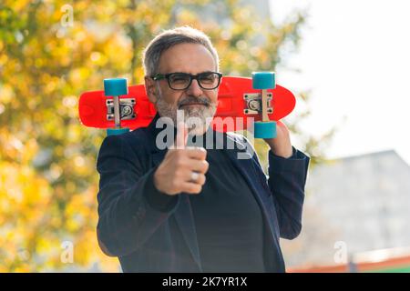 Grey-haired middle-aged man in glasses wearing blazer holding colorful skateboard and showing thumbs up to camera smiling. Silly grandpa. Horizontal outdoor shot. High quality photo Stock Photo