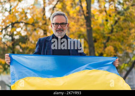 Grey-haired middle-aged man standing in city park holding Ukrainian flag looking into camera smiling. Supporting Ukrainians. Protest. Horizontal outdoor shot. High quality photo Stock Photo
