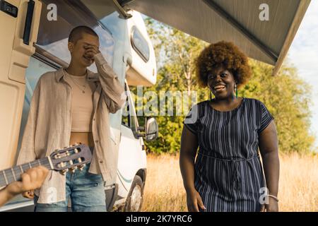 Acoustic guitar - live music during private party. Best friends - caucasian queer bald person and smiling Black woman in dark-blue dress - joking around and laughing in front of travel van. High quality photo Stock Photo