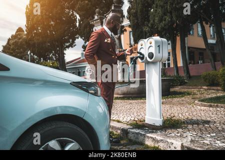 Side view of a dapper bald bearded black man in an elegant tailored chestnut-color costume checking the socket connection, a plug, and a power cable l Stock Photo