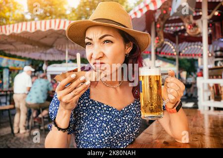 A girl in a pub sniffs a spoiled currywurst sausage and makes a disgruntled face while holding a beer in her other hand Stock Photo