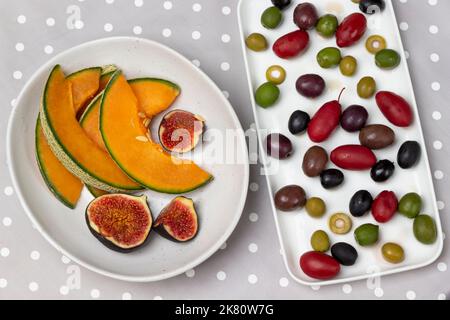 Olives of various varieties on plate, melon slices and cut figs in bowl. Flat lay. Gray polka dot background. Stock Photo