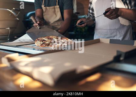One hot pizza, coming right up. two unrecognizable men standing and preparing a freshly made pizza for takeaway in their restaurant. Stock Photo