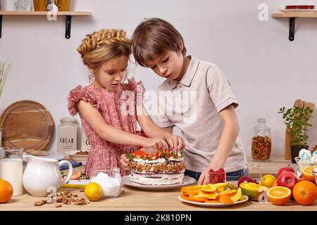 Little friends are making a cake together at a kitchen against a white wall with shelves on it. Stock Photo