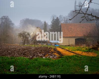 plowed field covered with a snow and old farmhouse on a background in an old village in a rainy misty winter day Stock Photo