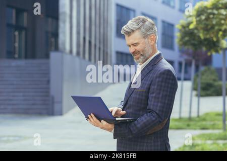 Happy and smiling boss using laptop while standing outside modern office building, mature businessman in business suit, senior investor satisfied with work result. Stock Photo