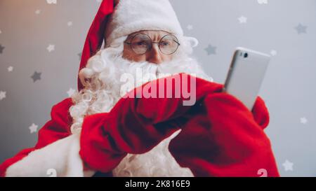 Serious guy in Santa outfit is using smartphone touching screen standing alone on grey starry background. Modern devices and communication concept. Stock Photo