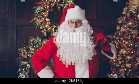 Portrait of Santa holding bag of gifts near Christmas tree in decorated house looking at camera with kind face. Man is wearing traditional red costume and glasses. Stock Photo