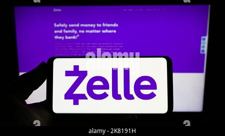 Person holding smartphone with logo of US digital payments company Zelle on screen in front of website. Focus on phone display. Stock Photo