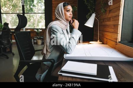 Thoughtful Muslim architect working with blueprints in a modern office. Innovative young businesswoman planning a new design project. Creative busines Stock Photo