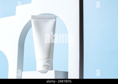 White unbranded plastic squeeze tube for cream or shampoo stands on white podium in white arch on blue background. Cosmetics showcase. Mockup Stock Photo