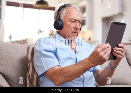 Excuse me whos there. an elderly man wearing headphones and using a digital tablet on the sofa at home. Stock Photo