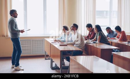 Male professor in casual clothes is talking to group of students sitting at tables in classroom and making notes. Large lecture hall with desks, chairs and large windows is visible. Stock Photo