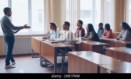 Male professor in casual clothes is talking to group of students sitting at tables in classroom and making notes. Large lecture hall with desks, chairs and large windows is visible. Stock Photo