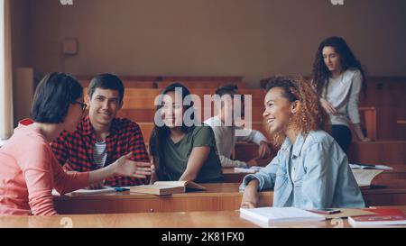 Careless girls and boys are talking and laughing sitting at desks in lecture hall, students are relaxing during break. Conversation, modern education, fun and youth concept. Stock Photo