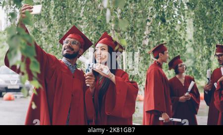 Two students are taking selfie with graduation diplomas wearing mortarboards and gowns, guy is holding smartphone and taking picture, girl is posing. Stock Photo
