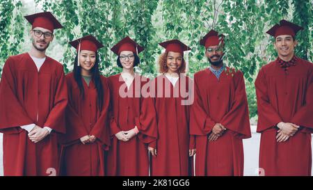 Portrait of multiethnic group of graduating students standing outdoors wearing red gowns and mortar-boards, smiling and looking at camera. Youth and education concept. Stock Photo