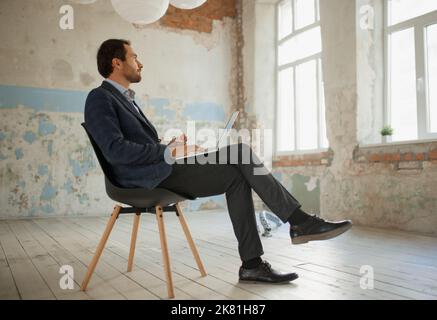Portrait of young businessman sitting on chair in empty room and working on laptop, texting messages. Business failure Stock Photo