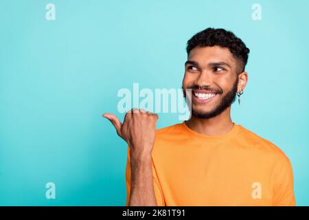 Photo of funky dreamy man dressed orange t-shirt looking pointing thumb empty space isolated turquoise color background Stock Photo