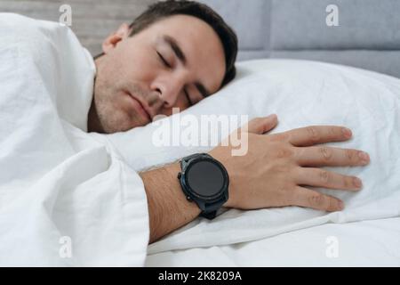Young man's head lying on white pillow in bed. Modern, black smart watch on wrist. Place for digital numbers, logo or mobile app on empty screen. hear Stock Photo