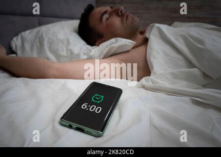 Man Sleeping Through Alarm Clock On Phone Lying In Bed At Home. Smartphone User Holding Cellphone While He Sleeps. alarm clock on a smartphone near a Stock Photo