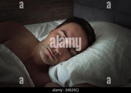 Handsome young man sleeping in white bedding. Head close-up on a pillow while sleeping at night. Stock Photo