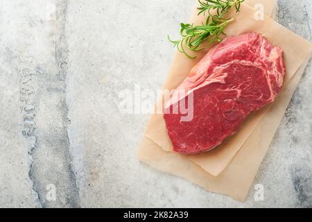Raw beef steak. Fresh beef rib eye steak with fork rosemary, salt and pepper on piece of parchment paper on dark grey stone background. Top view. Mock Stock Photo