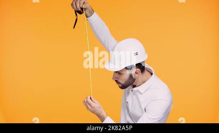concentrated architect in safety helmet looking at measuring tape isolated on yellow Stock Photo