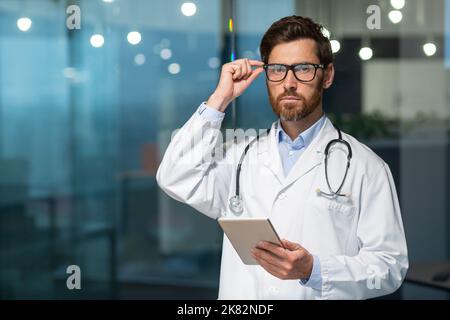 Portrait of a mature doctor with a beard, the man is looking at the camera in glasses and holding a tablet computer in his hands, the doctor is working inside the office of a modern clinic. Stock Photo