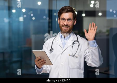 Senior mature doctor with tablet looking at camera and holding hand up greeting gesture, man in white medical coat working inside modern clinic, using tablet computer at work. Stock Photo