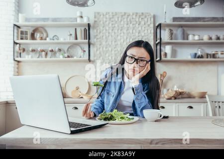 Dissatisfied and tired of dieting young Asian woman. He is sitting at the table in the kitchen, watching a nutritionist's webinar on a laptop. He holds a fresh salad, looks into the camera. Stock Photo