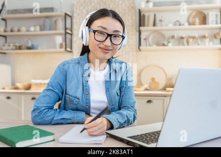 Young beautiful Asian woman working as a freelancer. Sitting at home with a laptop and wearing white headphones, writing in a notebook, looking at the camera, smiling. Stock Photo