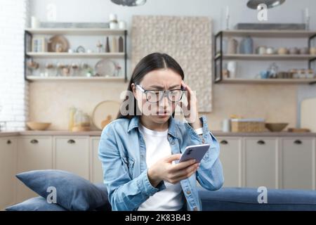 Worried young Asian woman holds the phone in her hands, received bad news by message. Sitting at home on the sofa, holding her head, nervous. Stock Photo