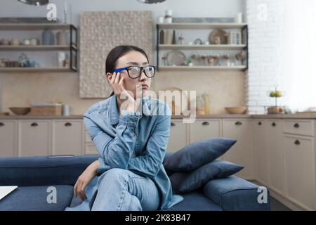 Depression. Pensive young beautiful Asian woman sitting on the sofa at home, looking out the window, resting her head on her hand, having a bad mood, anxious thoughts. Stock Photo